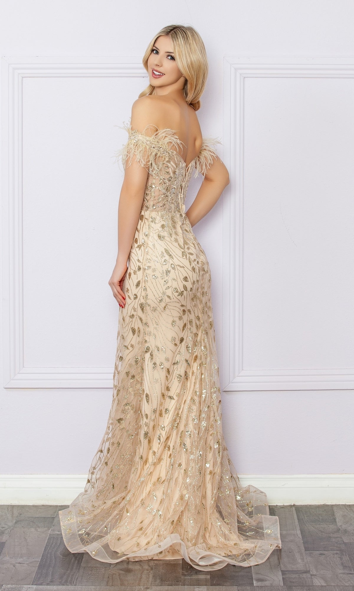 Woman wears the Nox Anabel L1255 Sequined Glitter-Print Prom Dress, featuring a light gold, off-the-shoulder design with sequin and feather accents, standing and looking over her shoulder against a pale wall.